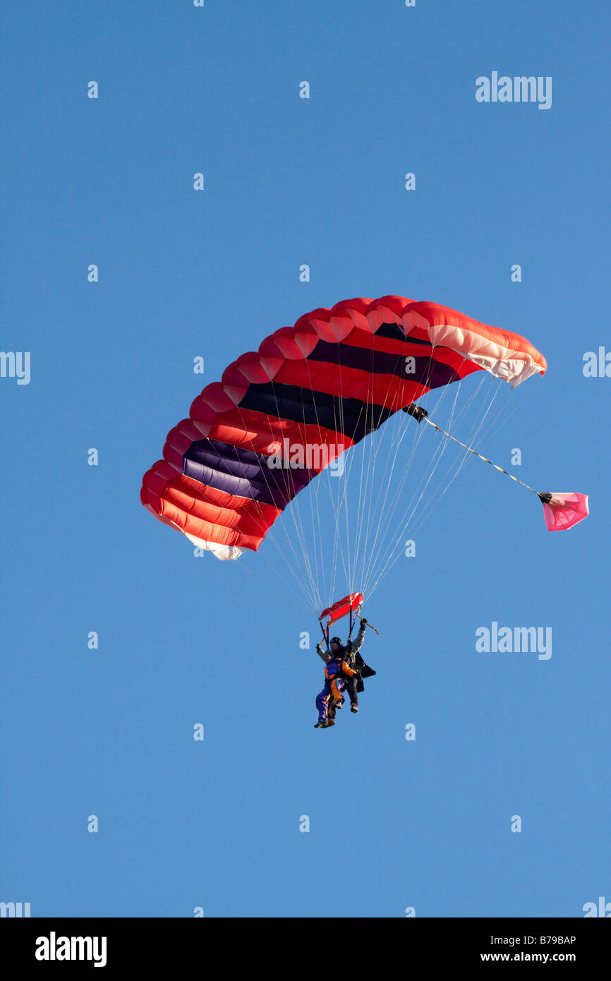 TANDEM PARACHUTING IN ENGLAND A RED STRIPED PARACHUTE CARRYING TWO PEOPLE GLIDES ACROSS THE BLUE SKY Stock Photo