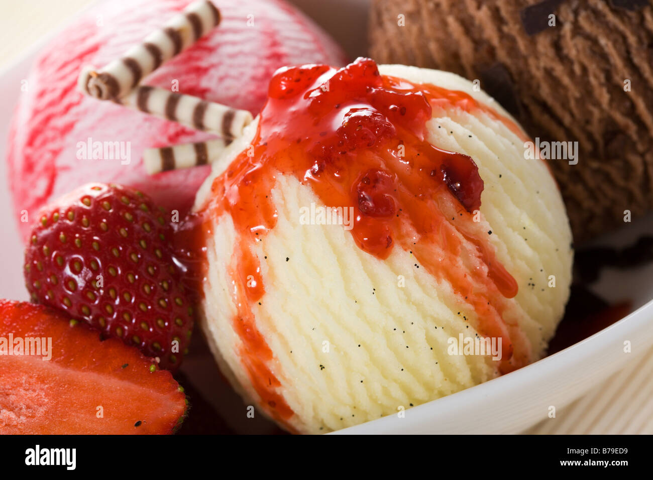 close-up of three balls of icecream - vanilla, strawberry, chocolate - with strawberry topping, strawberries and chocolate rolls Stock Photo