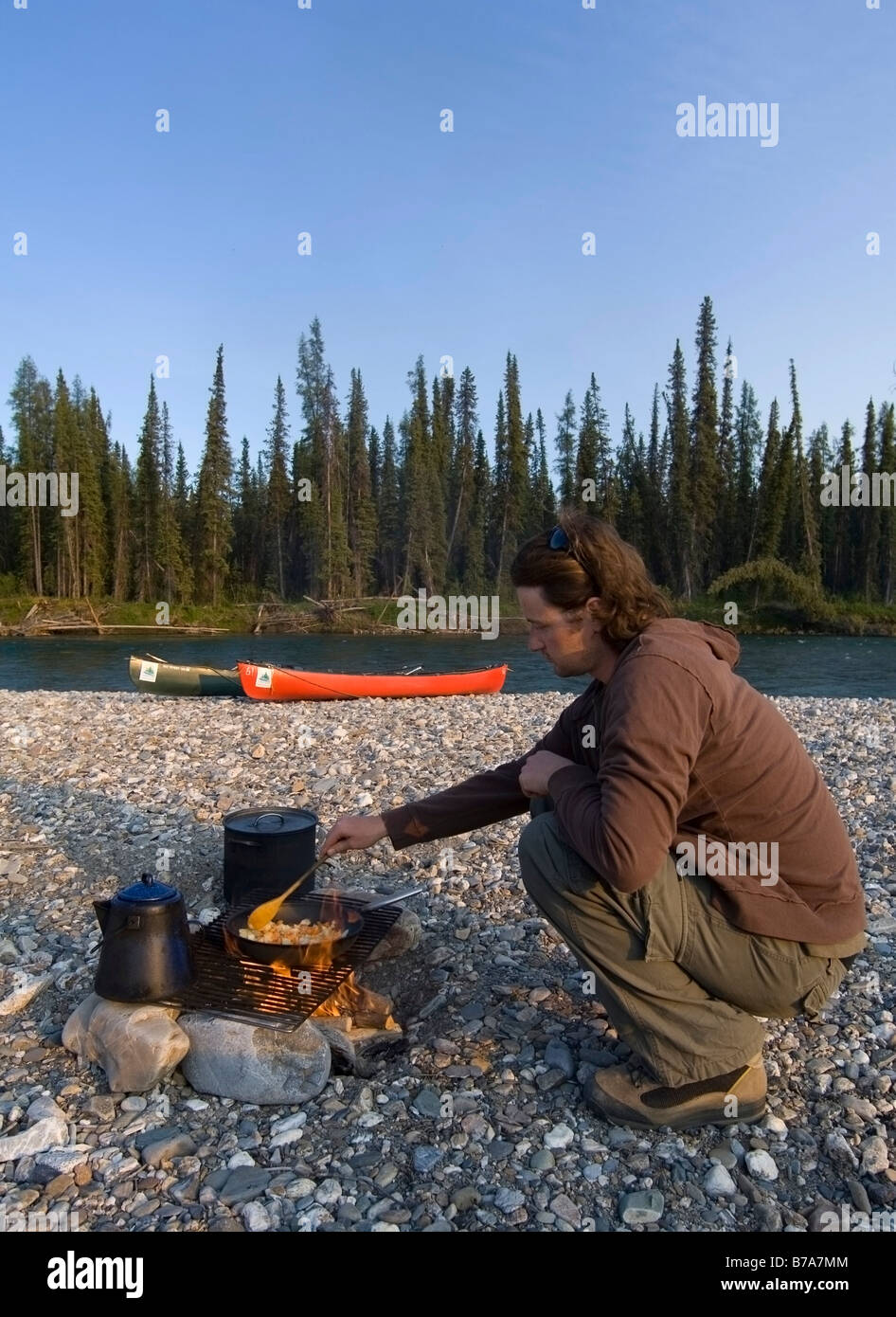 Man cooking on a camp fire, canoes behind, Liard River, Yukon Territory
