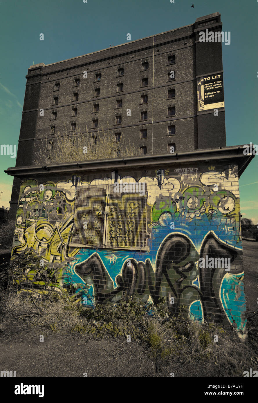 Cyan tinted and toned view of a customs bonded warehouse with small graffiti covered building in the foreground. Stock Photo