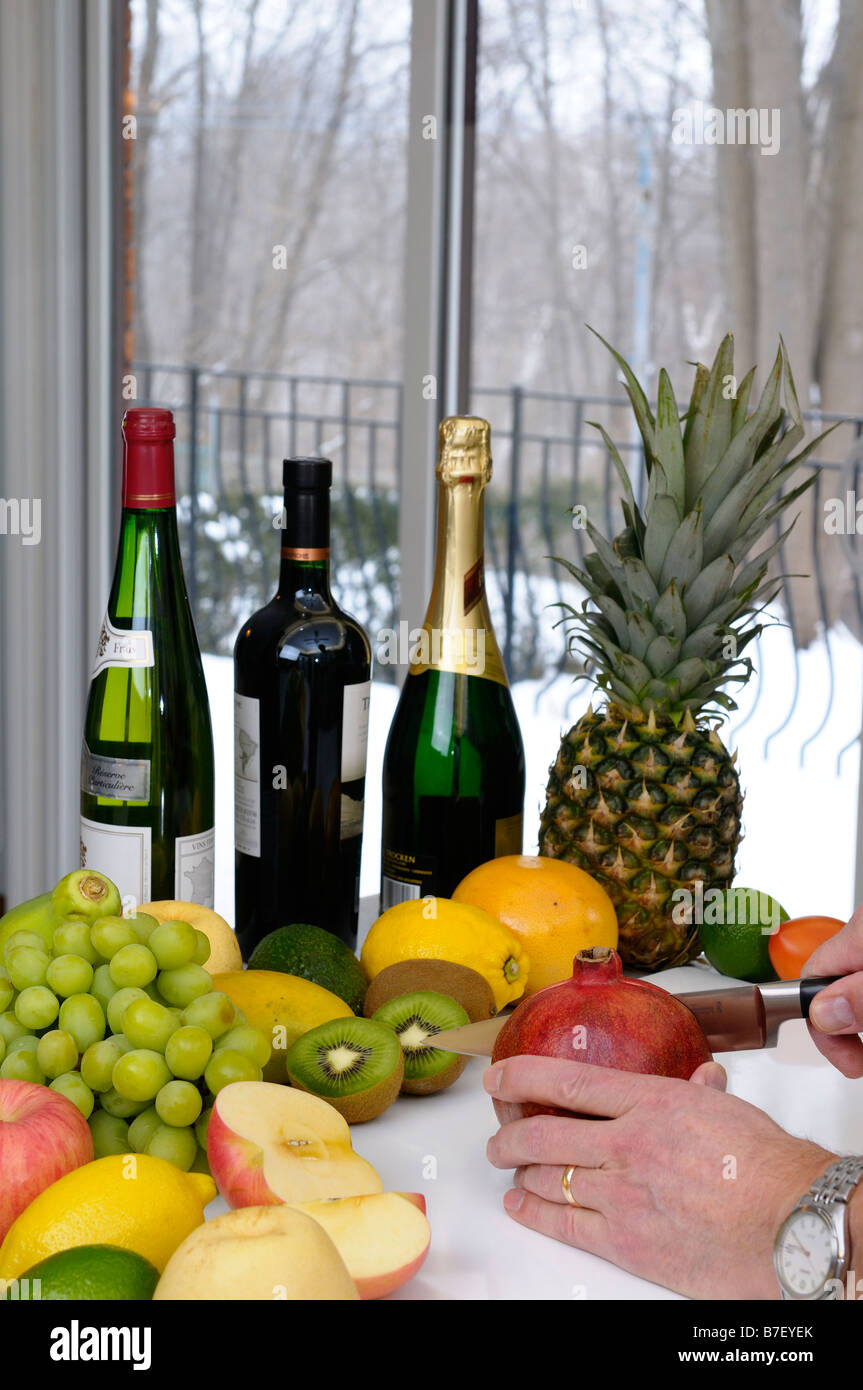 Man cutting into a pomegranate surrounded by fruit and wine in a kitchen on a winter day Stock Photo