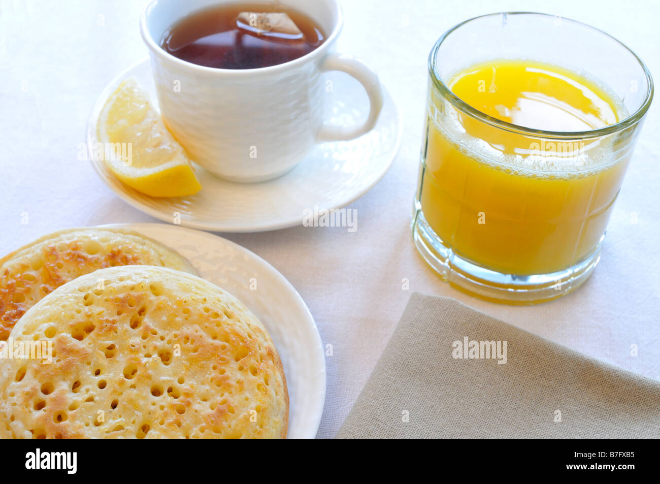 Crumpets on white plate with glass of orange juice, cup of black tea with lemon and napkin on white background. Stock Photo