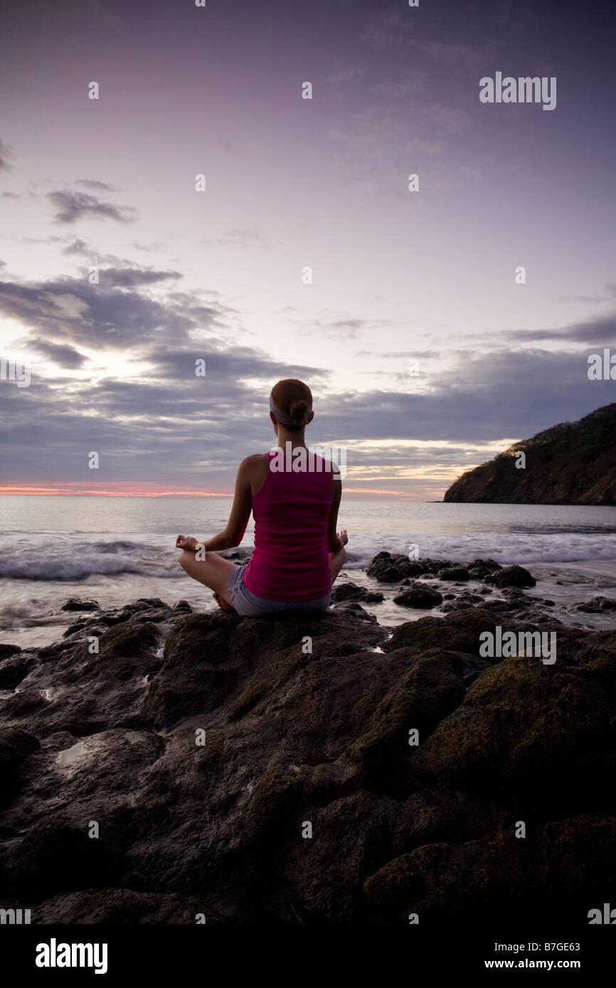 Young woman meditating on rocks at dusk in front of ocean surf in Playas del Coco, Costa Rica. Stock Photo