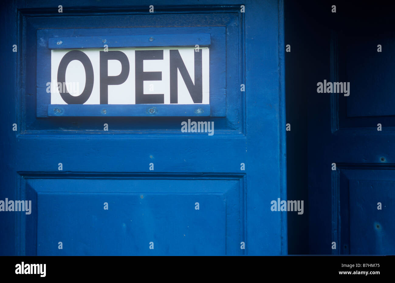 Black and white slide-in sign stating OPEN on dark blue painted panelled door with adjacent door standing ajar Stock Photo
