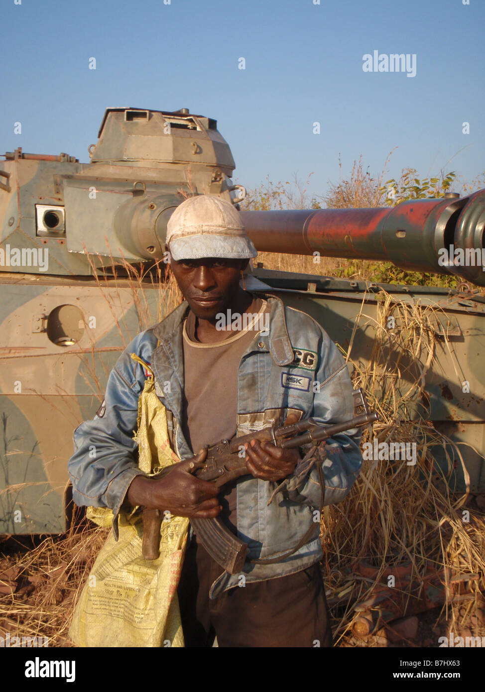 Armed Congolese man with AK47 automatic rifle with abandoned tank behind him near Lake Mweru Democratic Republic of Congo Stock Photo