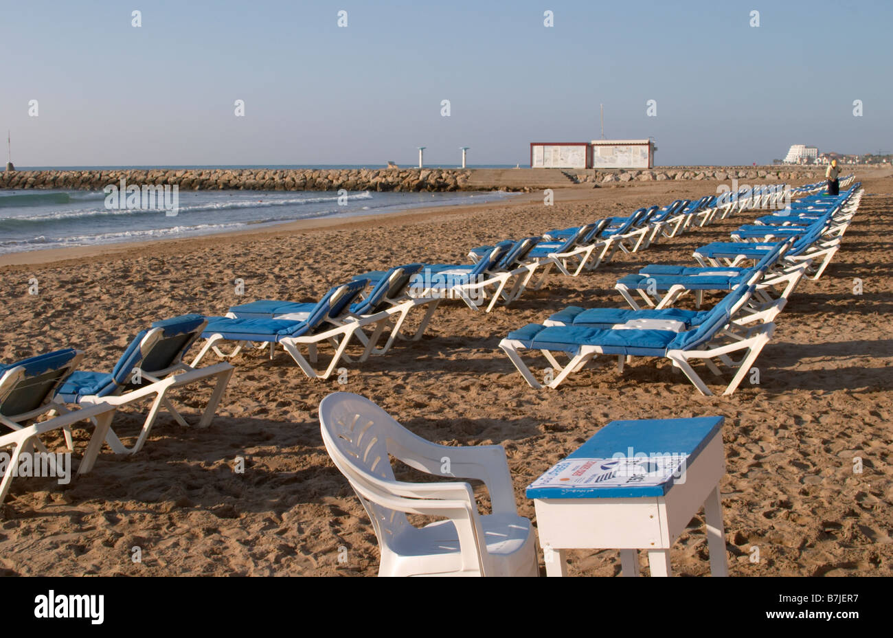 The beach. Rows of empty deck chairs. A plastic chair and parasol for