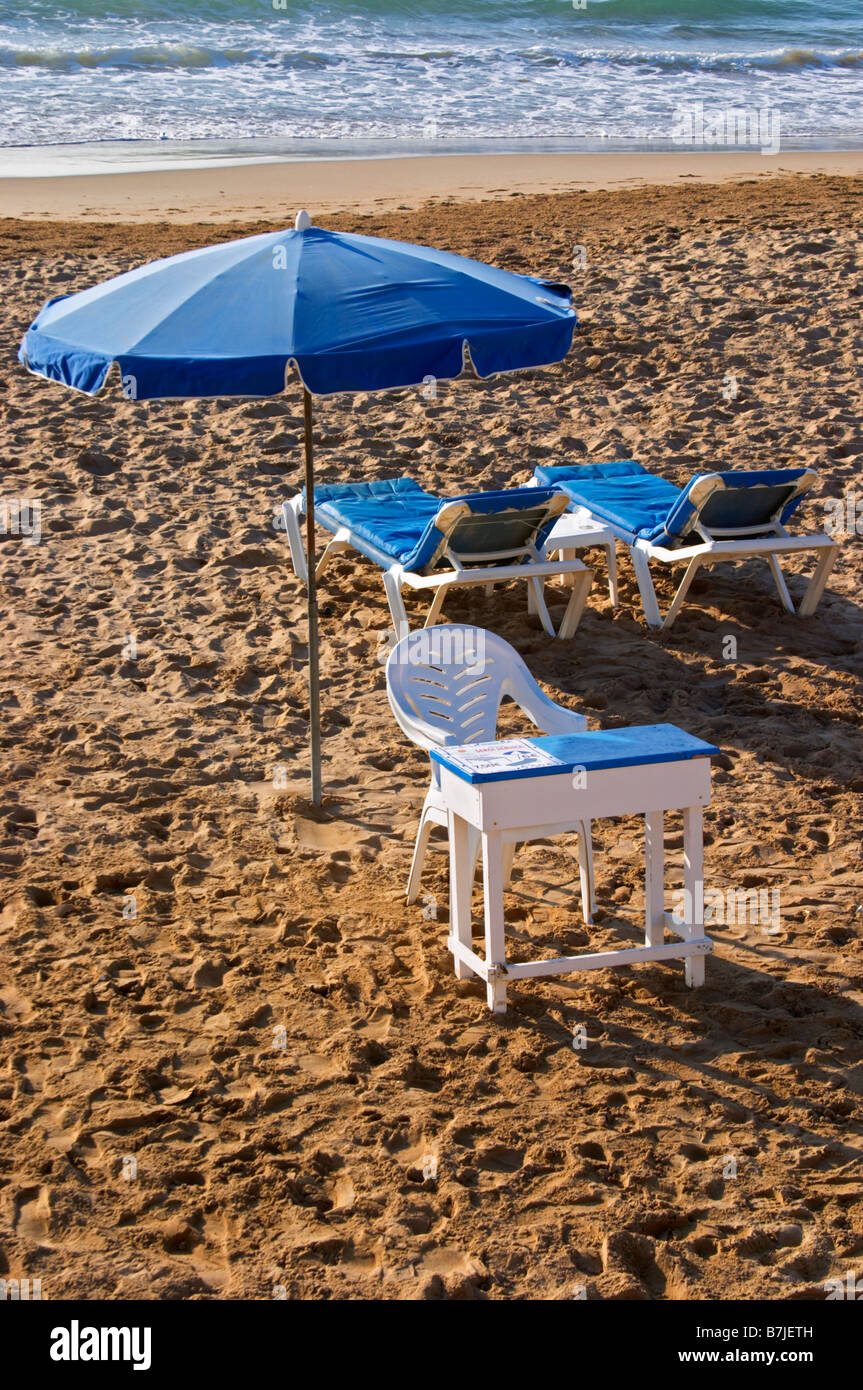 The beach. Two empty deck chairs and a parasol. A plastic chair and