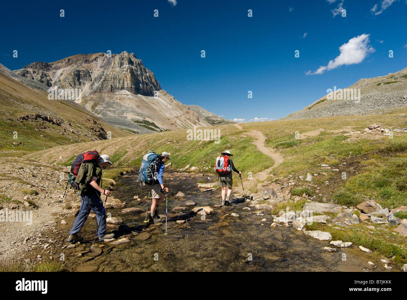 Three hikers cross a small creek in an alpine meadow.; Canada, Alberta, Jasper National Park, Skyline Trail Stock Photo