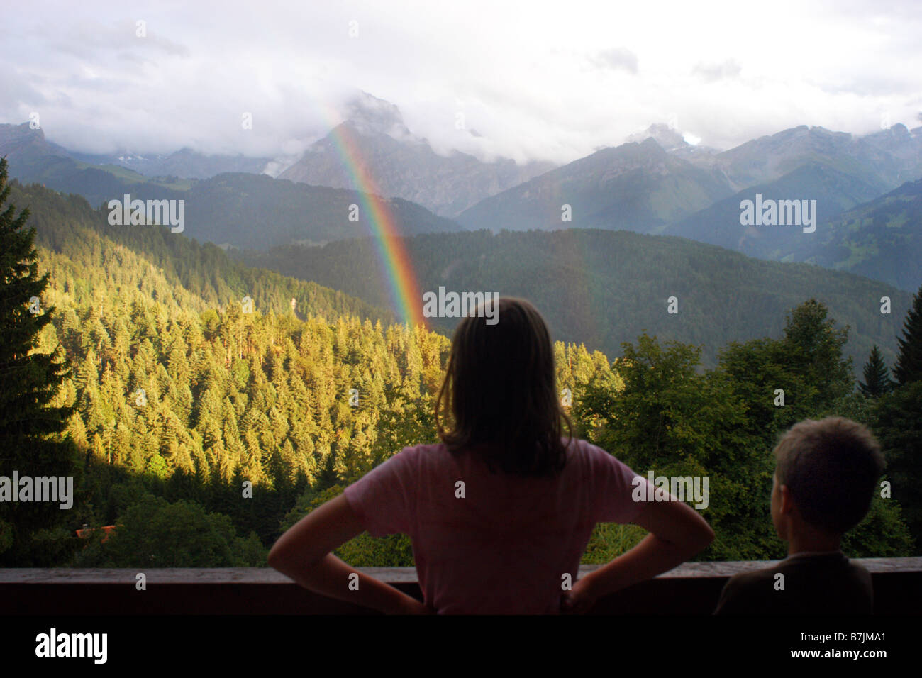 Two children look out of a swiss holiday chalet in Switzerland onto a rainbow and mountains Stock Photo