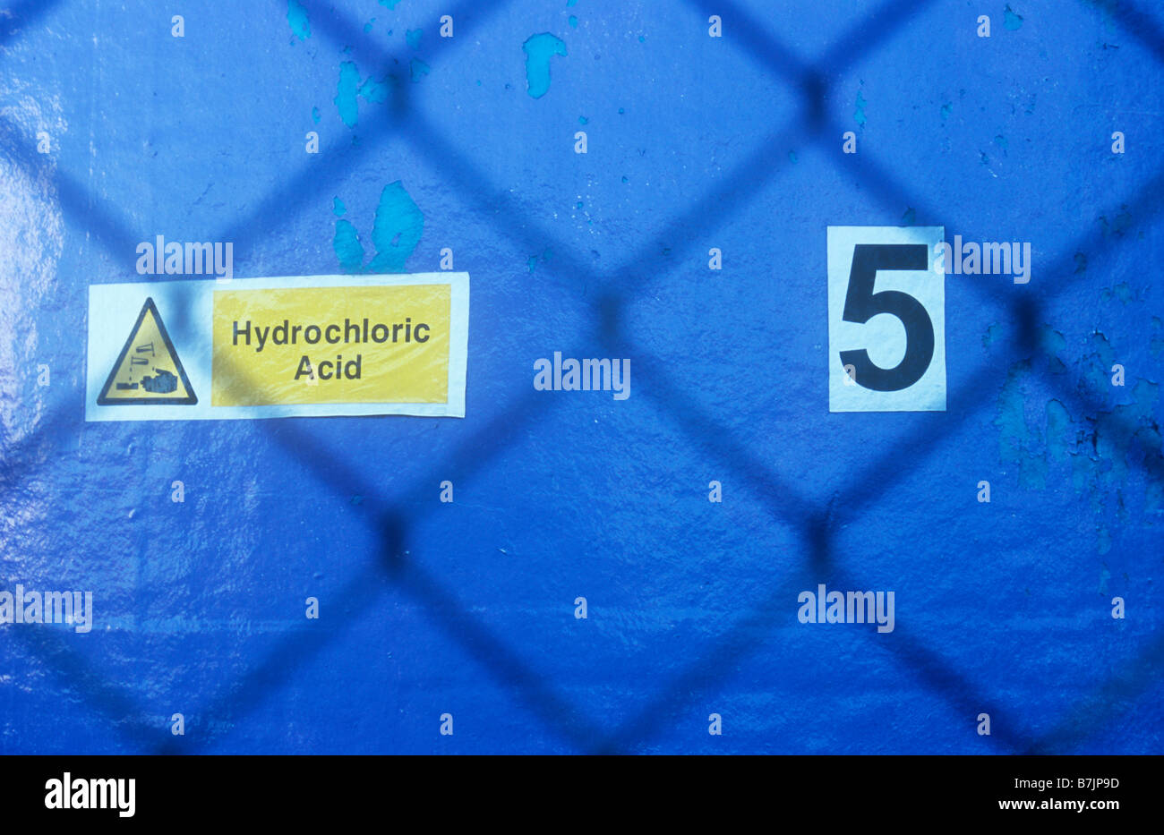 View through wire mesh security fence to violet-blue storage tank with signs stating 5 and Hydrochloric acid Stock Photo