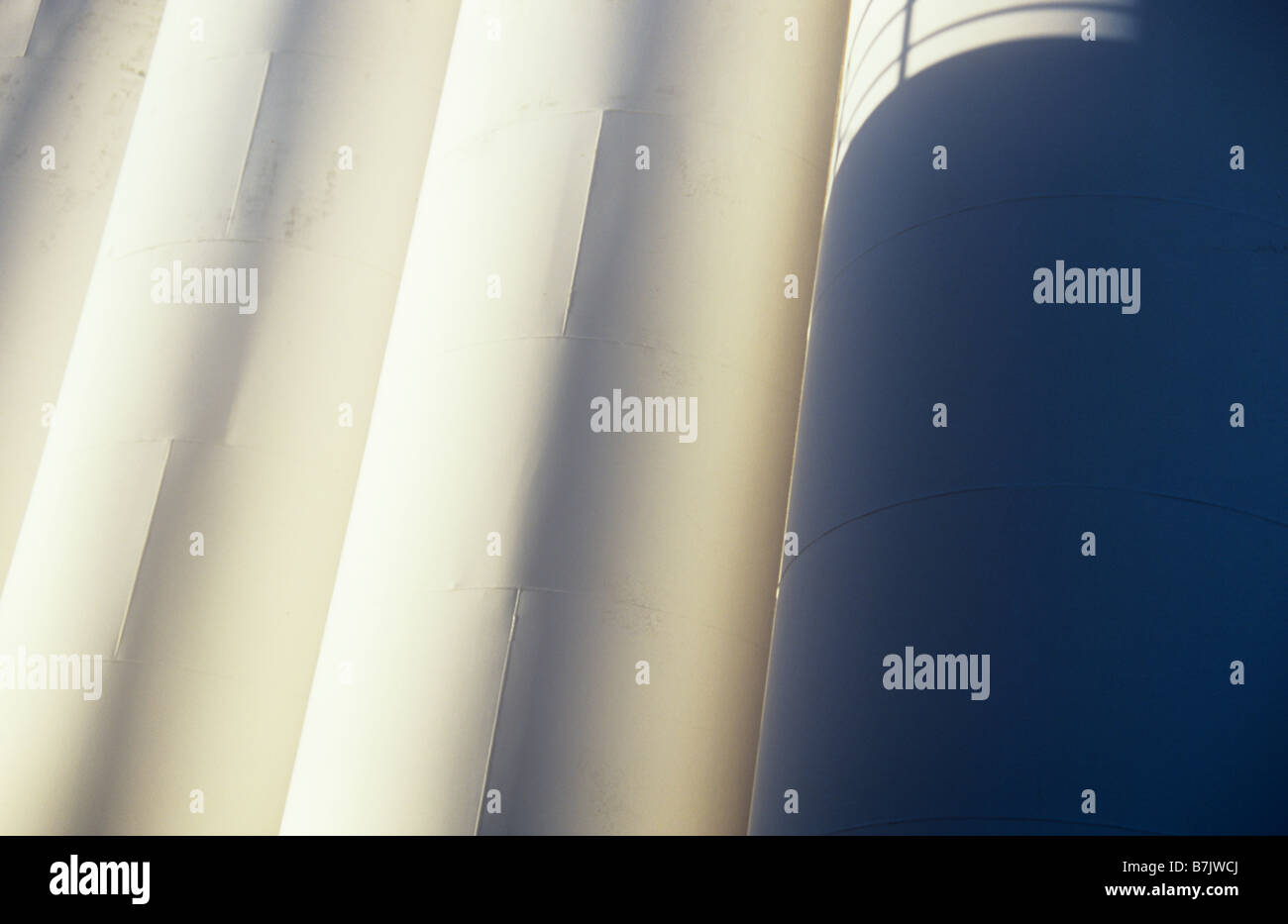 Low sunlight glancing across closely grouped white painted large industrial or petroleum storage tanks with shadow of railings Stock Photo