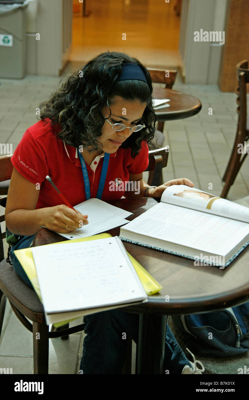 Female Hispanic college student sitting at library table writing notes from a textbook preparing and studying for a course. Stock Photo