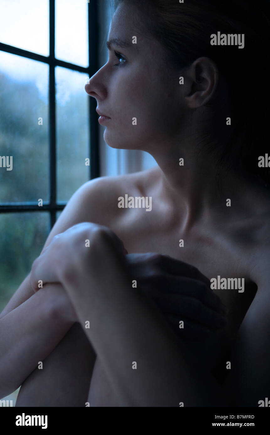 Thoughtful: a beautiful, pensive young woman (21+) sits naked by a window Stock Photo