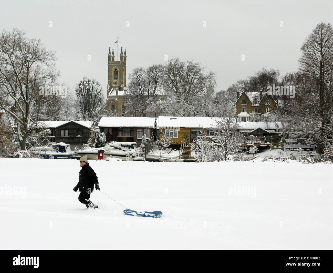 Boy pulls sled across riverside open space at Sadler's Ride, Hurst Park, East Molesey. Hampton church on other side of river in background. Stock Photo