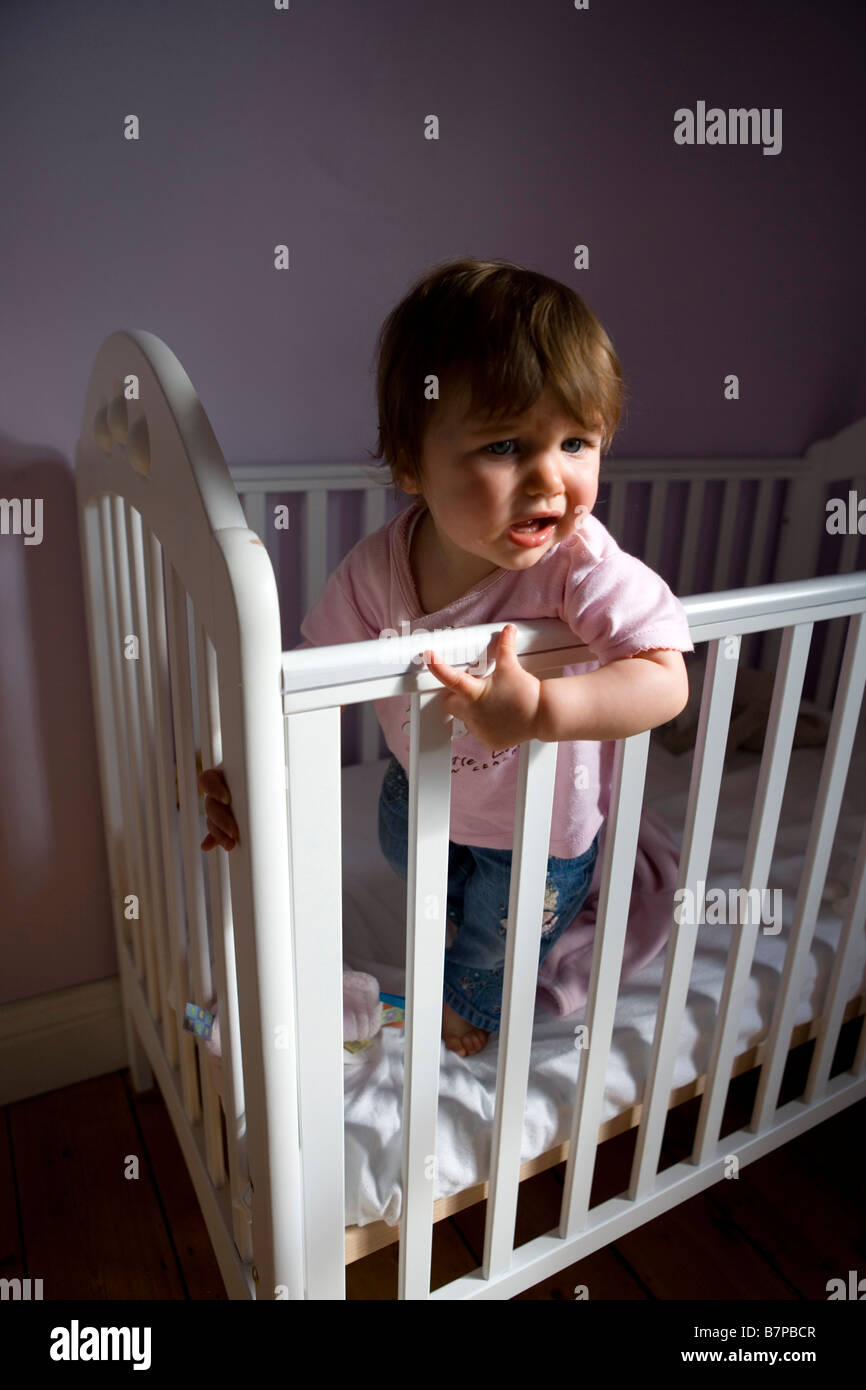 A unhappy looking  baby girl stands up in her cot . Stock Photo