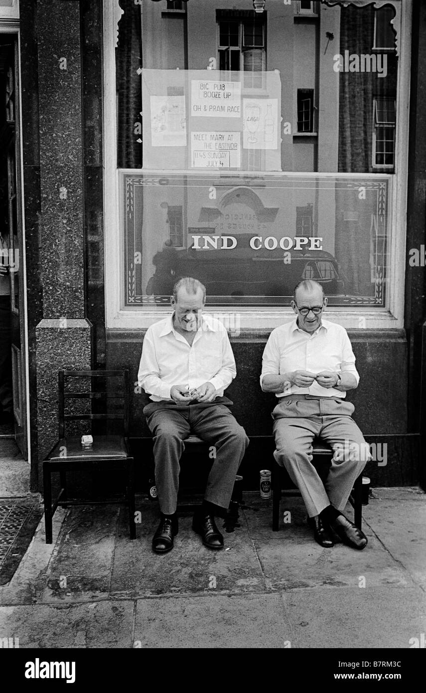 Two men in white shirts rolling cigarettes sitting outside their local pub. Somers Town London Stock Photo