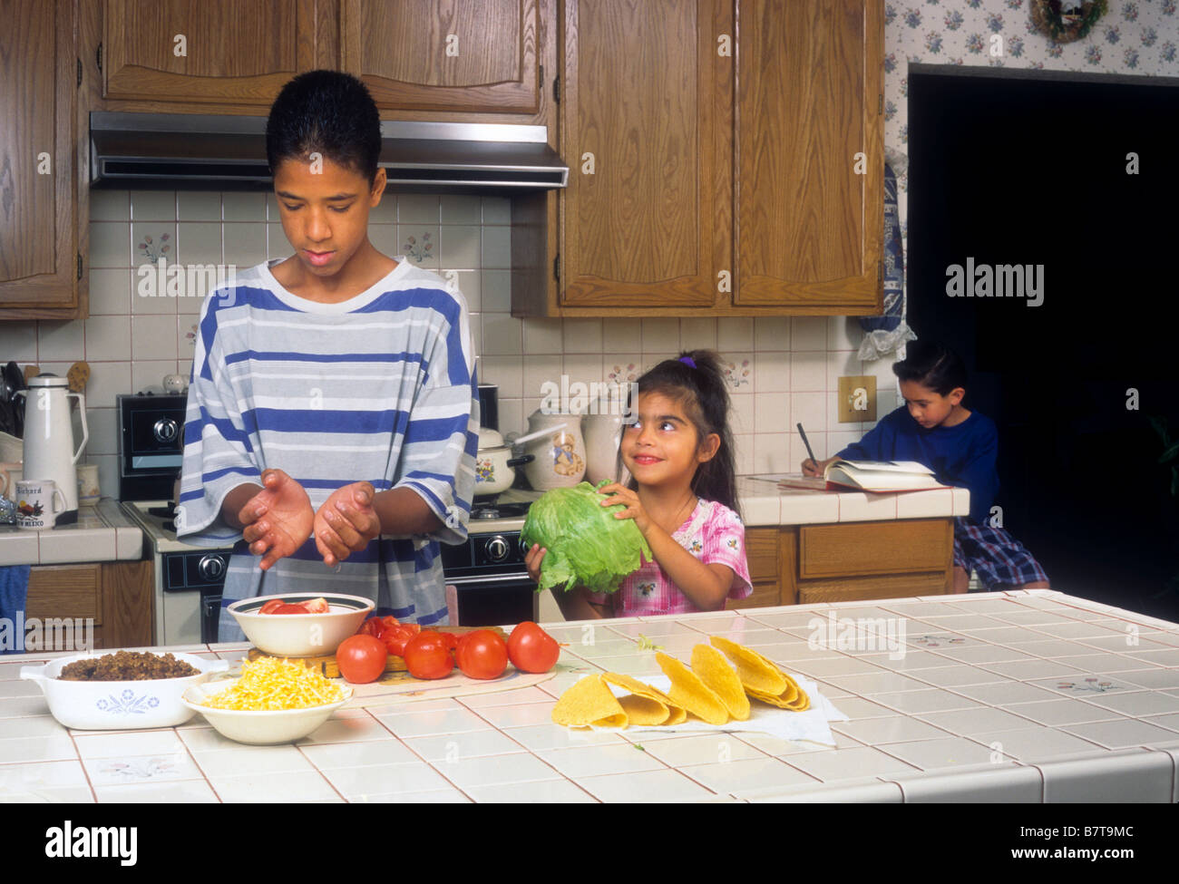 Little girl helps her older brother as he makes tacos for family as other brother does homework in background Stock Photo