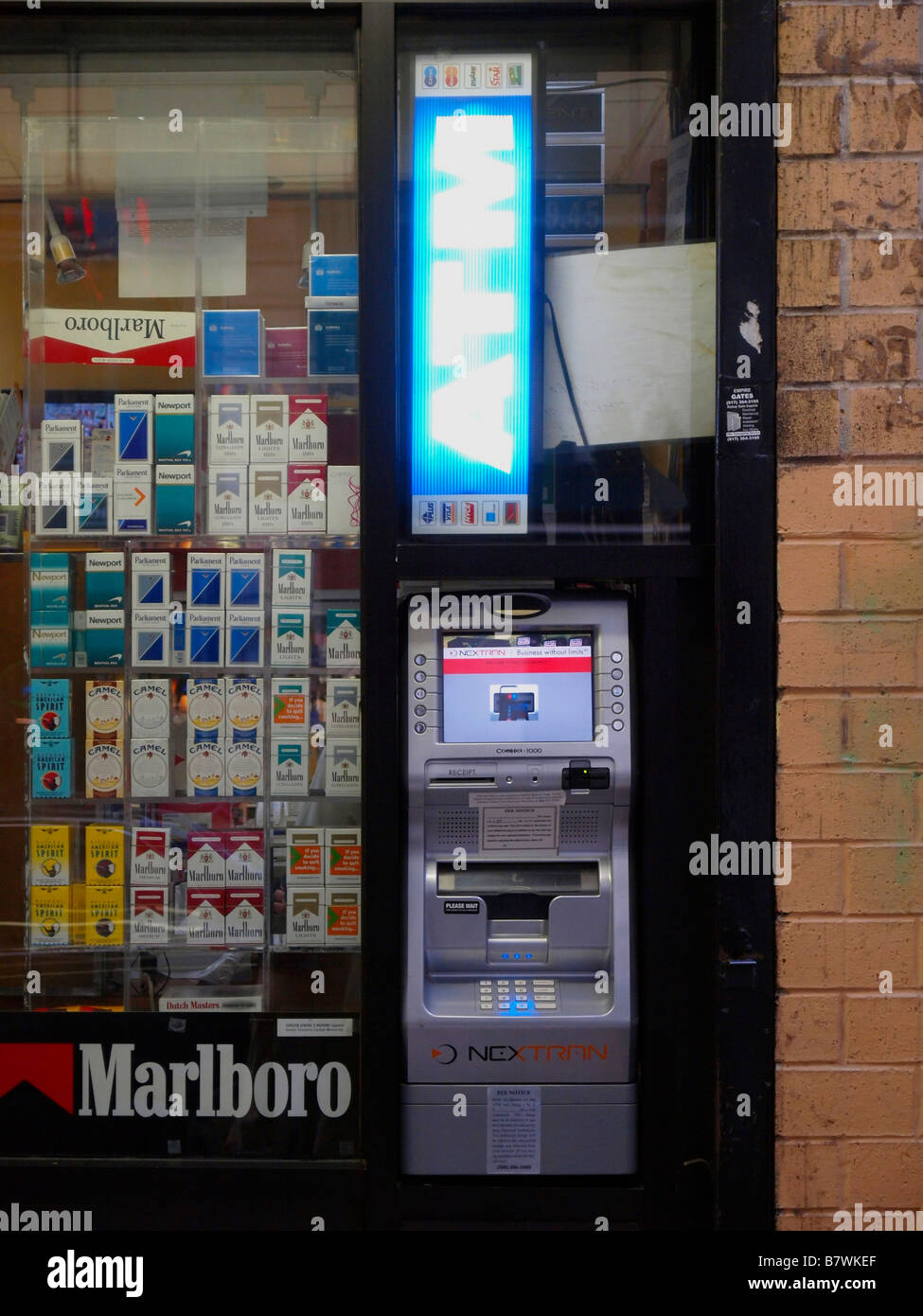 An outdoor automatic teller machine beneath a lighted 'ATM' sign next to a window display full of cigarettes in New York City. Stock Photo