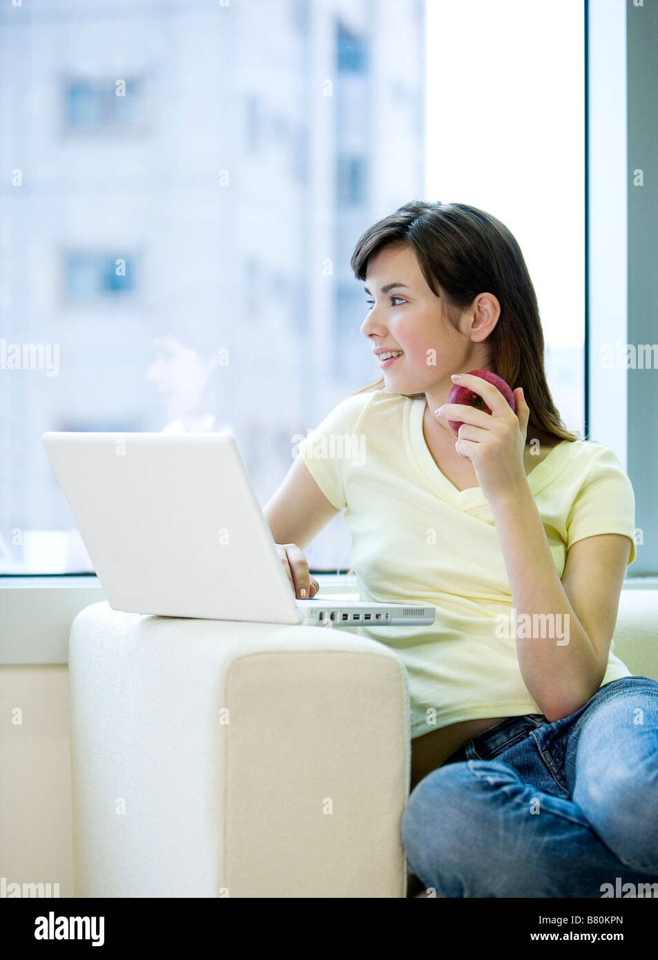 Young woman sitting on sofa with laptop holding an apple side view ...