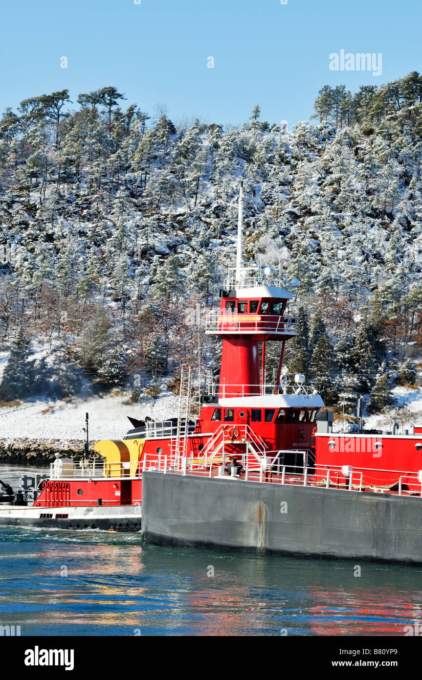 Scenic winter scene with tugboat pushing barge in water past Cape Cod coastline with trees covered in freshly fallen snow Stock Photo