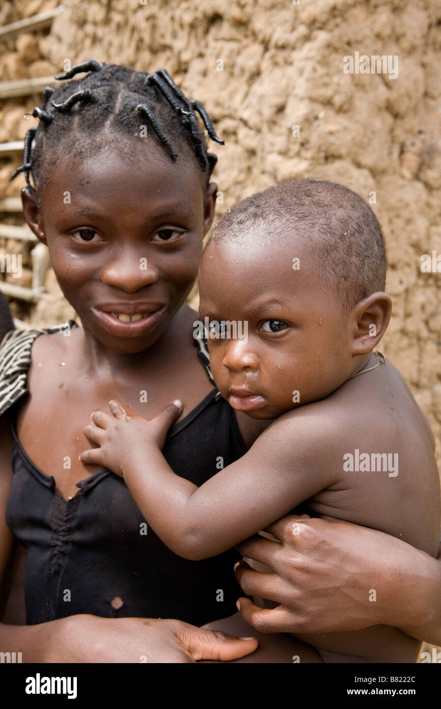 Young Nigerian girl holds her baby brother in the sun outside their wattle and daub mud hut of their remote jungle village Stock Photo