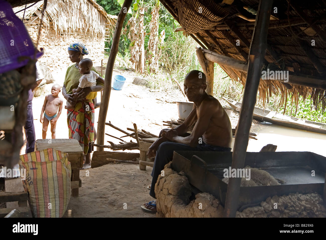 Old man from a remote Nigerian village sits smoking inside his home while the rest of his family stand outside in the sun Stock Photo