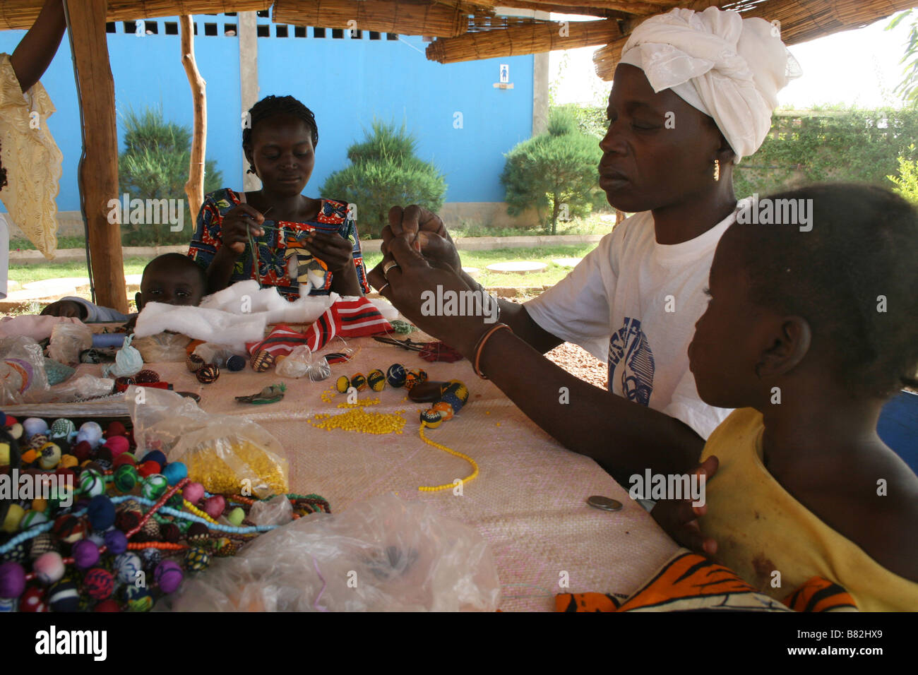 African craftwomen at work making fabric beads while their children watch Stock Photo