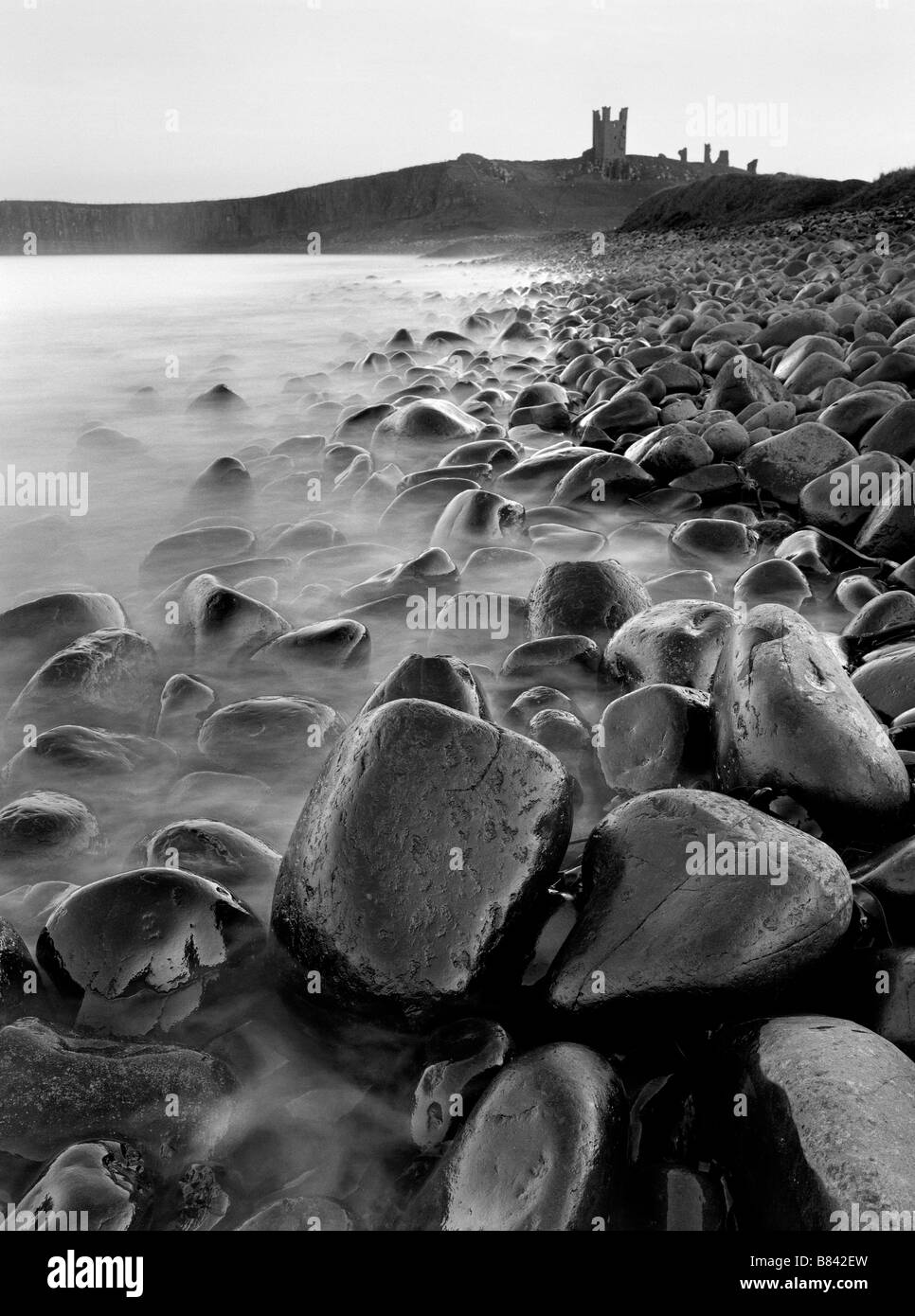 The waters lapping the rocks look like mist, on the beach at Embleton with a view of Dunstanburgh Castle, Northumberland, UK Stock Photo