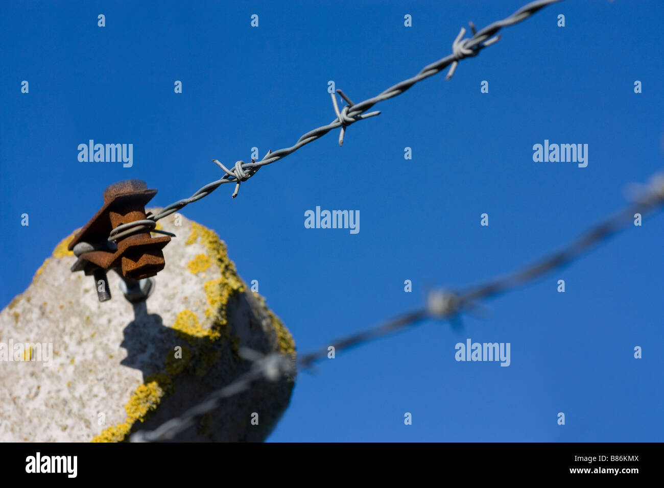 Tip of pre cast concrete security fence post with rusty barbed wire tensioner and 2 strips of barbed wire against blue sky Stock Photo
