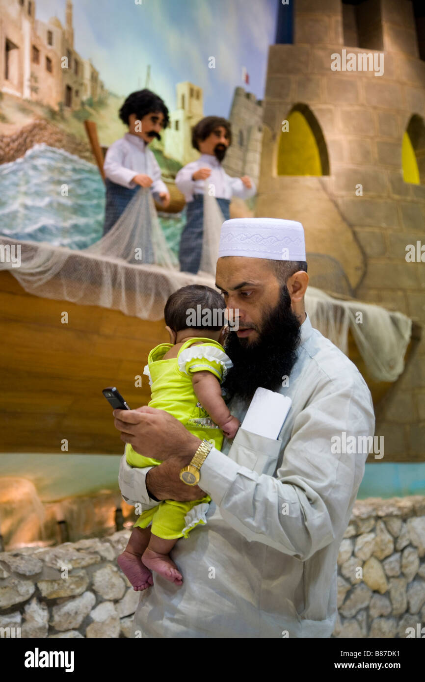 Muslim father texting and holding a baby, in front of three dimensional travel display in departure lounge of Bahrain airport. Stock Photo