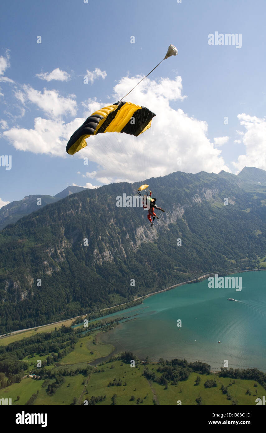 Skydive couple under canopy is flying along the lake side torts the mountain with clouds and blue sky. Stock Photo