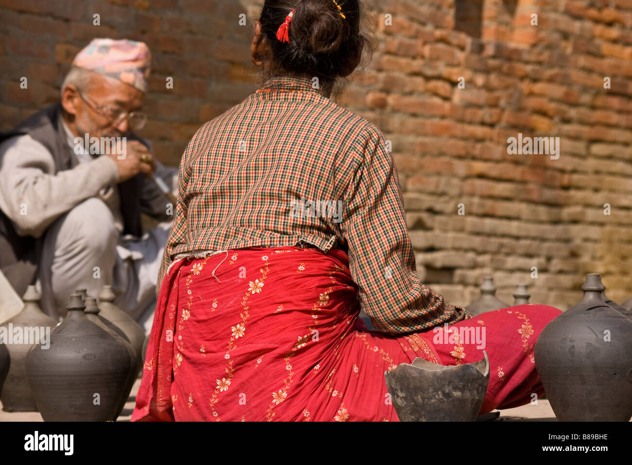 Old Nepali man and pot seller in traditional clothes with hat sits smoking hookah pipe with woman outdoors on the street in Bhaktapur old town, Nepal Stock Photo
