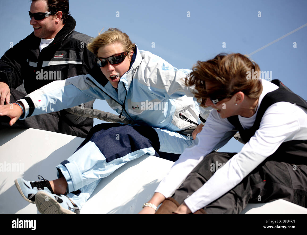 3 adults,2 woman and 1 man leaning over the edge of a yacht while racing. The picture is color in a landscape format. Stock Photo