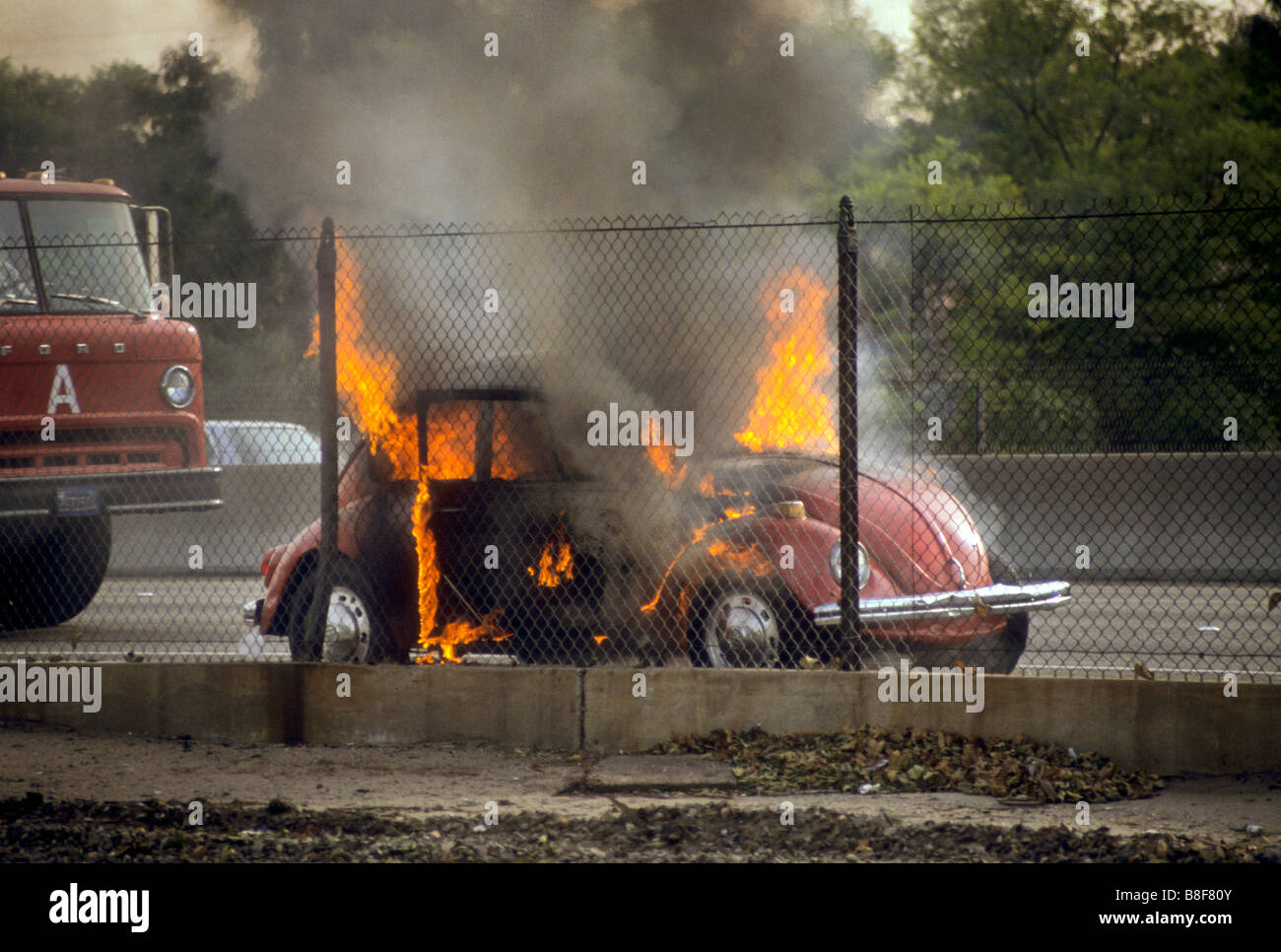Volkswagen bug auto sits in flames on side of freeway as tire truck arrives. Stock Photo