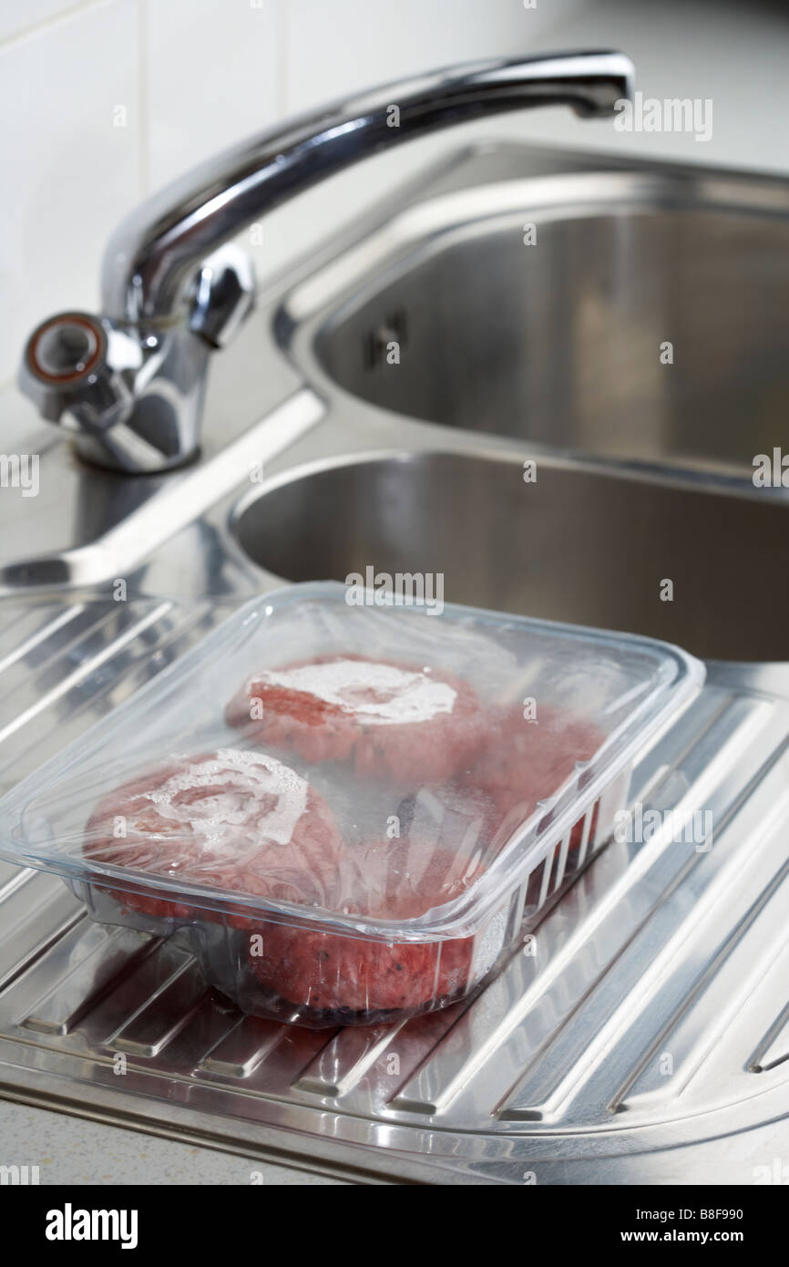 four beefburgers in a plastic tray defrosting at room temperature on a kitchen sink draining surface Stock Photo