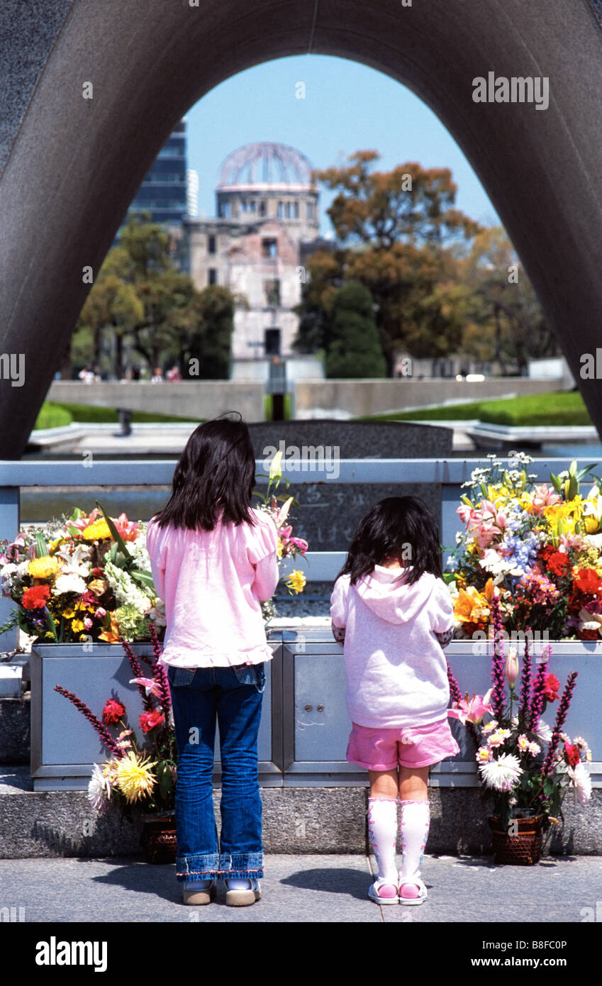 Two girls say a prayer in front of the Peace Memorial and in the distance the A Bomb Dome in Hiroshima Japan Stock Photo