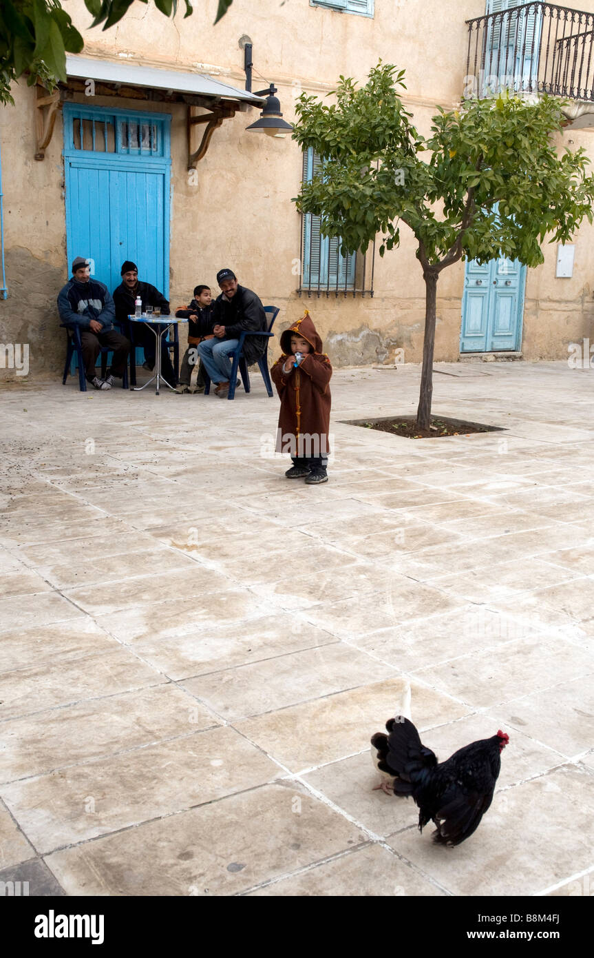 Outside a café in Testour, Tunisia, a male-only family group is amused by the playful little boy entertained by a cockerel Stock Photo