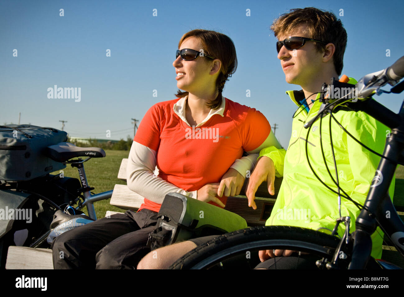 Young, attractive couple sit on a park bench next to their touring bicycles. Stock Photo