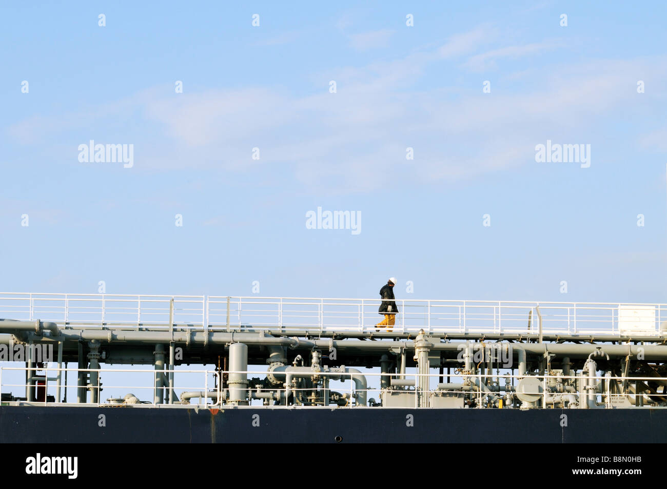 One man worker  walking across an industrial walkway with rails with pipes below. Stock Photo
