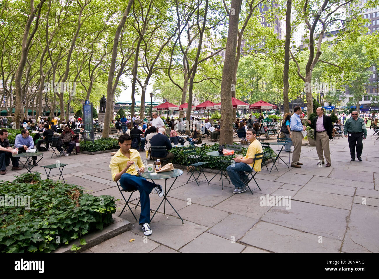People sitting at small tables under tall trees in a public garden Stock Photo