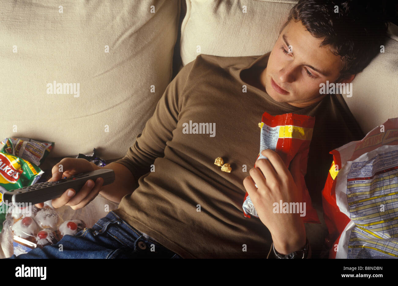 slobbish man lounging on a settee watching television and eating junk food Stock Photo