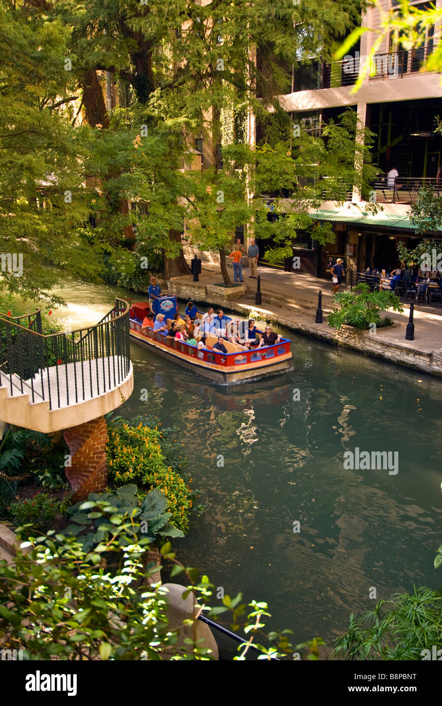 San Antonio River Walk riverwalk tour boat with tourists passes beside outdoor cafes and trees with bright green leaves Stock Photo