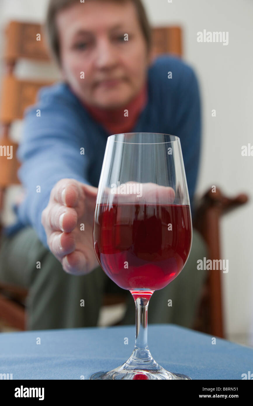 A mature woman alone in need of a drink sitting down reaching to pick up glass of red wine on a table in front. England UK Britain Stock Photo