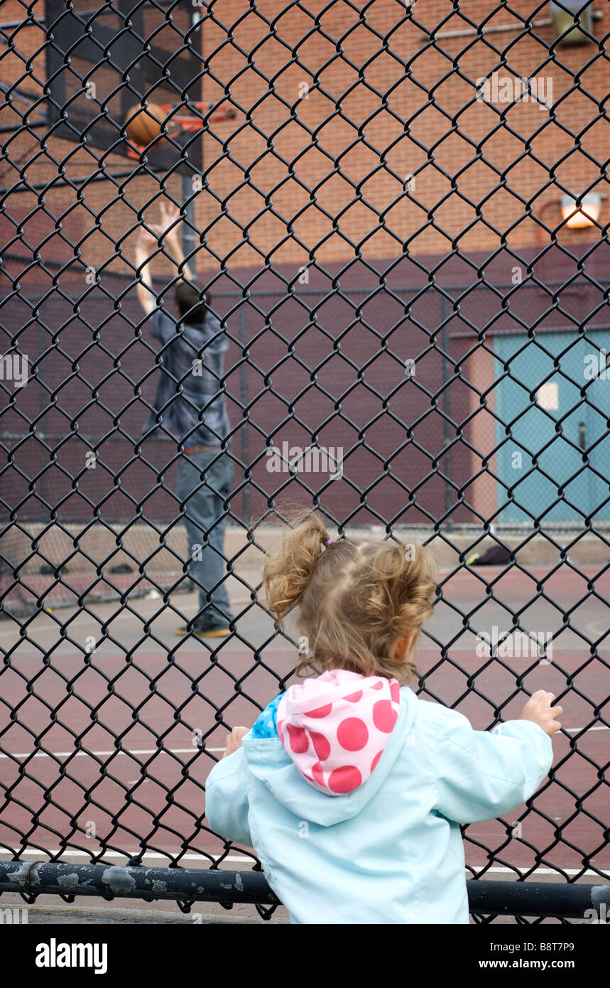 Little Girl in Pigtails Stands At Fence, Watching Older Teen Play Basketball in School Playground Stock Photo