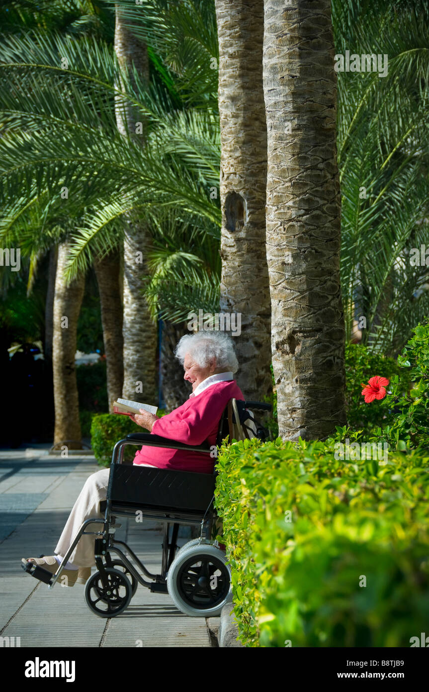 Elderly disabled lady on vacation wheelchair garden traffic free zone sitting reading a book in sunny sunlit holiday safe lush green palm tree setting Stock Photo