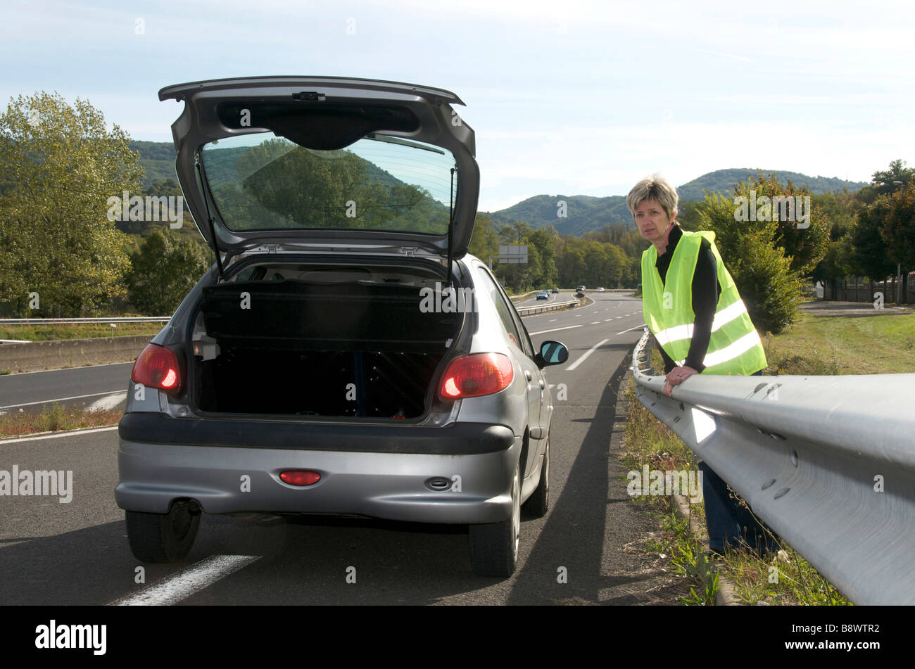 Woman standing behind the armco barrier with her broken down car waiting for help on a motorway slip road Stock Photo
