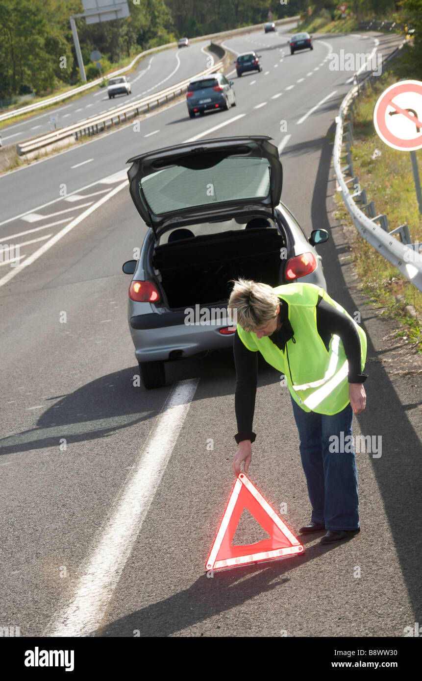 Woman wearing hi-viz placing a red warning triangle behind her broken down car on autoroute motorway slip road in France, Europe Stock Photo