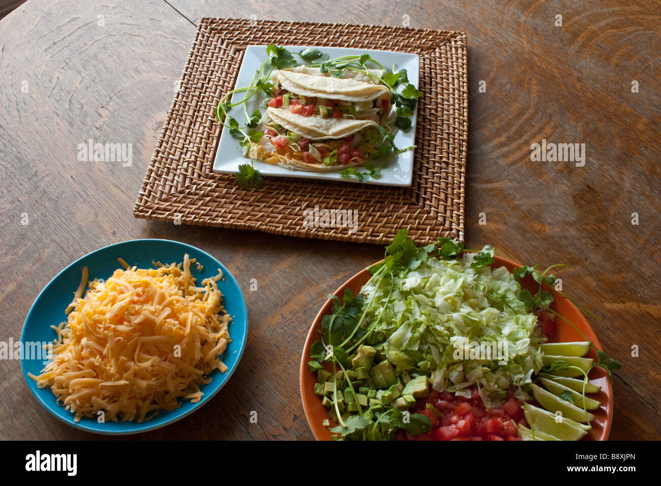 A plate of traditional style soft tacos on a bamboo charger garnished with cilantro and two platters of taco ingredients Stock Photo