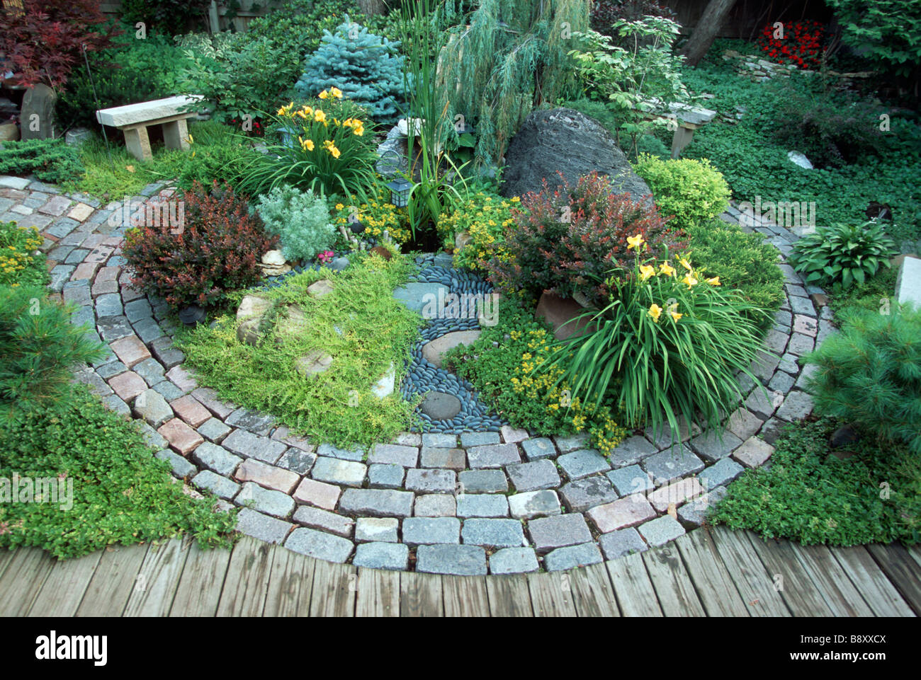 RECYCLED CITY STREET PAVERS AND BRICKS FORM PATTERN IN MINNESOTA PERENNIAL GARDEN.  BARBERRY, LILIES, GROUNDCOVERS AND BENCH. Stock Photo