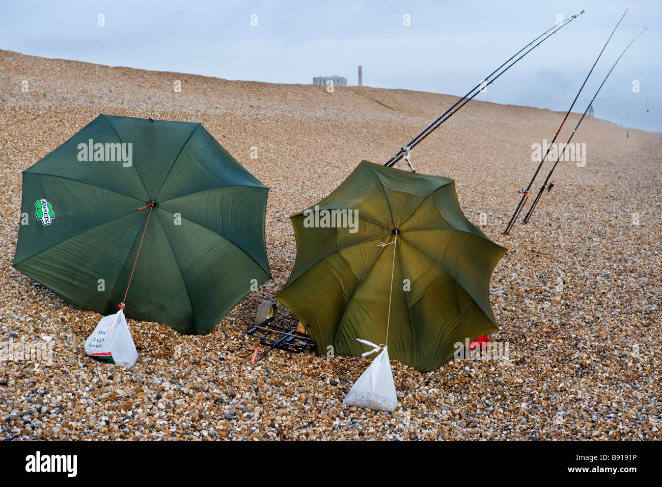 Two sea anglers fishing from the shingle beach using their umbrellas as windbreaks on Dengemarsh Beach, Dungeness in Kent, UK Stock Photo